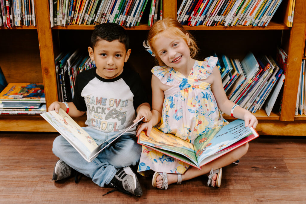 Two children holding a book while seating in front of a library bookshelf