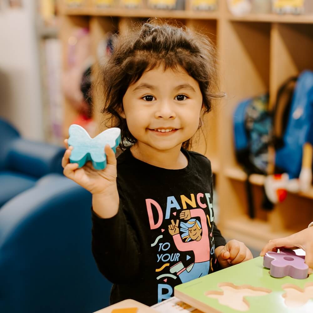 A cheerful girl holding a butterfly puzzle piece, symbolizing creativity and play in early childhood education.